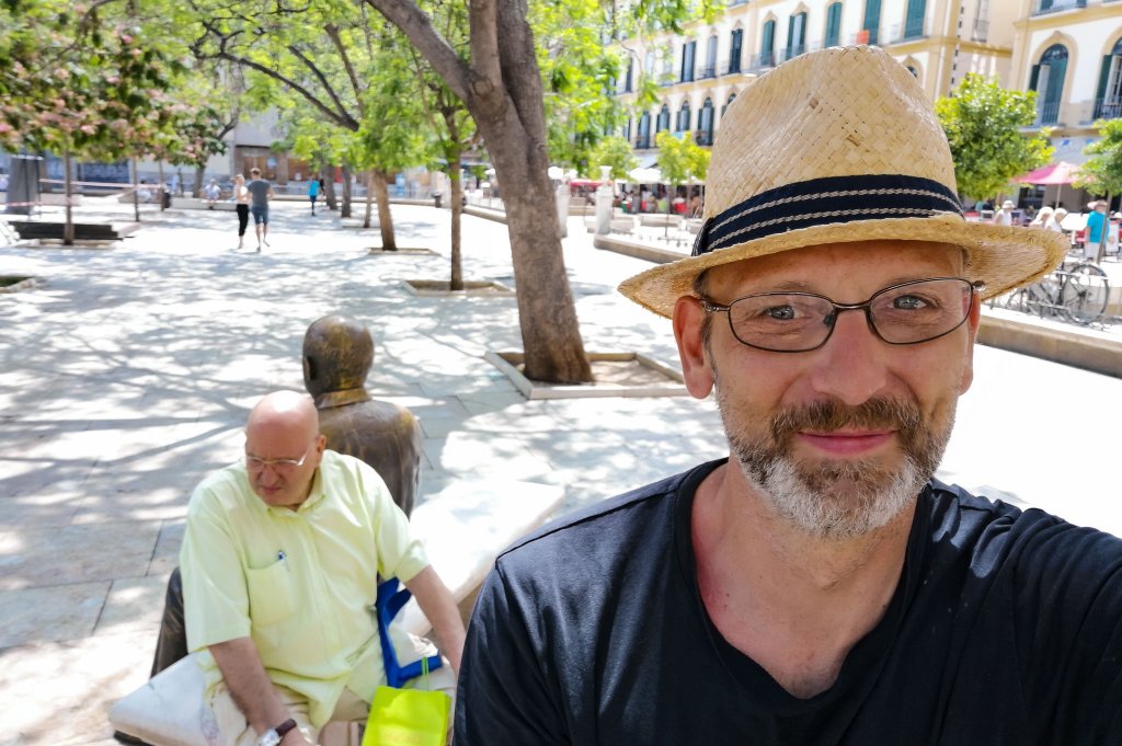 Selfie with straw boater, Picasso statue with a local sitting beside him. in the backgound.