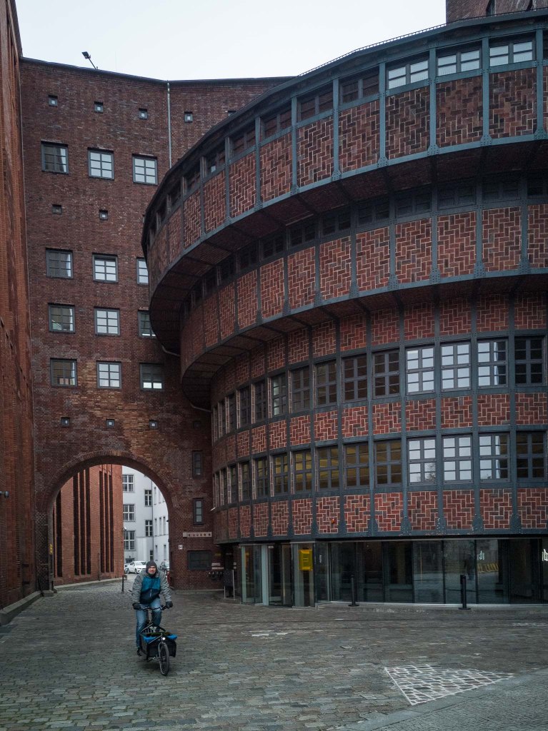 Dunkle, mit Klinker verkleidete Gebäude mit einer Durchfahrt, durch die gerade ein Mann auf einem Lastenrad fährt. Der rechte , runde Gebäudeteil wird in Stufen nach oben hin breiter.