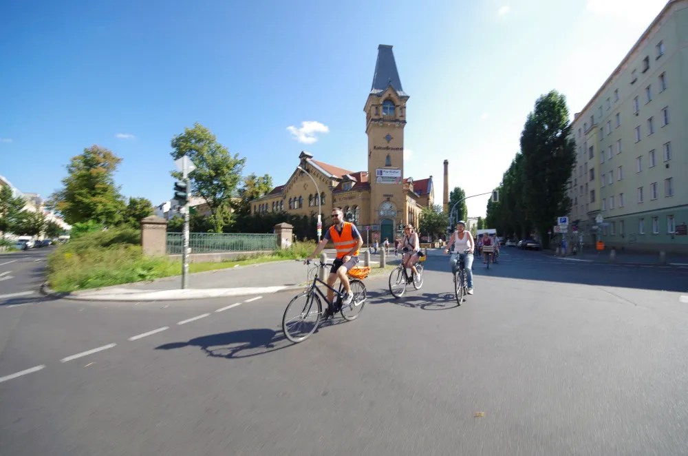 Cyclists ride down a sunny street with a historic building and trees in the background.