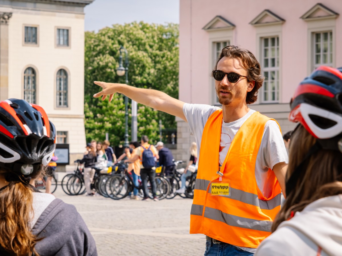 Tour guide in orange vest points while cyclists listen, wearing helmets.