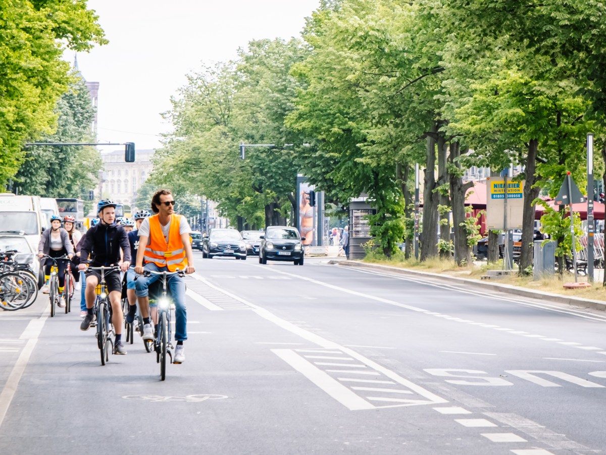 Cyclists in a city bike lane, led by a person in an orange vest, with cars on the road beside them.