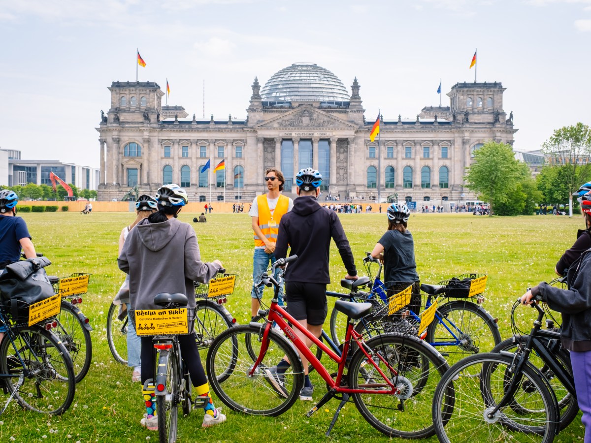 Cyclists gather near a historic building with a glass dome and flags.